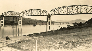 Photo is taken from the bank of a river and shows a bridge under construction with two people on a bike watching from the shore. The bridge consists of multiple iron spans on high concrete pylons. Fields are seen on the bank behind the bridge. 