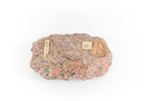 A small, solid granite specimen with a relatively flat top. The colour consists of flecks of pink, red, and grey.  It has three sticker tags.