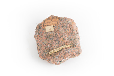 A small, solid granite specimen with a relatively flat top and right side. The colour consists of flecks of pink, red, and grey.  It has three sticker tags (see transcription).