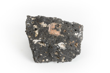 A smaller than hand sized rock specimen which is dark grey in colour with small holes and white deposits across its surface.