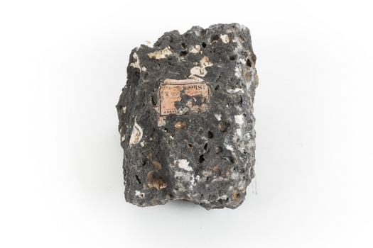 A smaller than hand sized rock specimen which is dark grey in colour with small holes and white deposits across its surface.