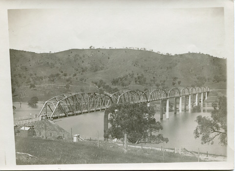 A bridge over a river featuring 9 arches. Background is rolling hills spotted with trees. In the foreground there is wire fence running parallel to the river.