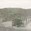 A bridge over a river featuring 9 arches. Background is rolling hills spotted with trees. In the foreground there is wire fence running parallel to the river. 