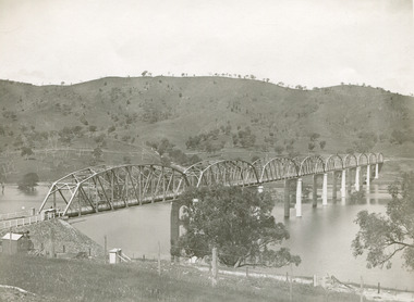 A bridge over a river featuring 9 arches. Background is rolling hills spotted with trees. In the foreground there is wire fence running parallel to the river. 
