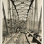 A bridge under construction in sepia. The perspective is from inside the bridge, looking along to one end, with iron trusses on either side, iron struts and bracing above, and floor beams below strewn with wooden planks.