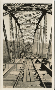 A bridge under construction in sepia. The perspective is from inside the bridge, looking along to one end, with iron trusses on either side, iron struts and bracing above, and floor beams below strewn with wooden planks.