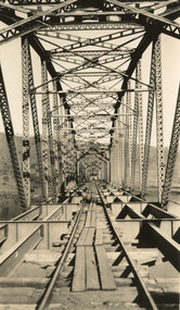 A bridge under construction in sepia. The perspective is from inside the bridge, looking along to one end, with iron trusses on either side, iron struts and bracing above, and floor beams below strewn with wooden planks.