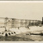 Partially constructed dam with scaffolding running along on what would later be the dam wall across the middle of the photograph, water pouring through pipes on the bottom third of photograph.
