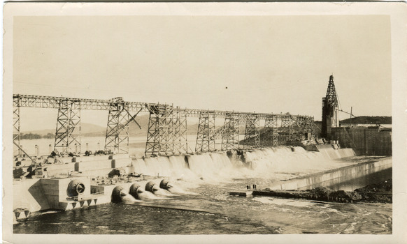 Partially constructed dam with scaffolding running along on what would later be the dam wall across the middle of the photograph, water pouring through pipes on the bottom third of photograph.