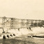 Partially constructed dam with scaffolding running along on what would later be the dam wall across the middle of the photograph, water pouring through pipes on the bottom third of photograph. 