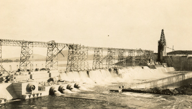 Partially constructed dam with scaffolding running along on what would later be the dam wall across the middle of the photograph, water pouring through pipes on the bottom third of photograph. 