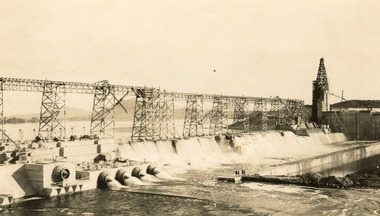 Partially constructed dam with scaffolding running along on what would later be the dam wall across the middle of the photograph, water pouring through pipes on the bottom third of photograph. 