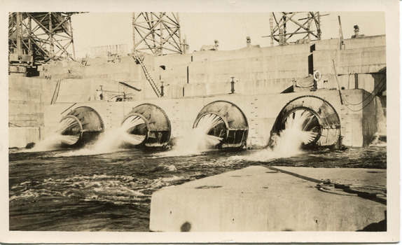 Open valves of a dam showing water flowing into stream of water, background shows scaffolding on wall of dam.