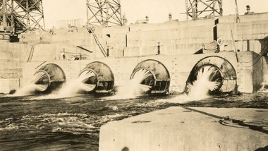 Open valves of a dam showing water flowing into stream of water, background shows scaffolding on wall of dam.