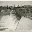 View of water flowing down the embankment of a dam to the left, with scaffolding running along on the top of the embankment. Also showing buildings in the background. 