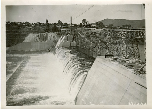 View of water flowing down the embankment of a dam to the left, with scaffolding running along on the top of the embankment. Also showing buildings in the background. 