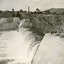 View of water flowing down the embankment of a dam to the left, with scaffolding running along on the top of the embankment. Also showing buildings in the background. 