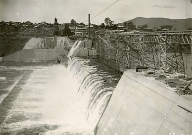 View of water flowing down the embankment of a dam to the left, with scaffolding running along on the top of the embankment. Also showing buildings in the background. 