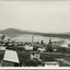 Many small cottages in the front half of the photograph, with a dam in the background, showing scaffolding for construction on the dam wall. Also showing the far bank of the dam with hills in the background.