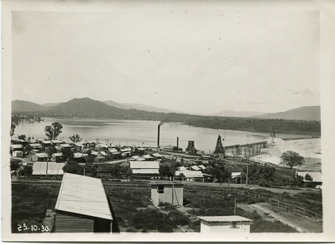 Many small cottages in the front half of the photograph, with a dam in the background, showing scaffolding for construction on the dam wall. Also showing the far bank of the dam with hills in the background.