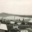 Many small cottages in the front half of the photograph, with a dam in the background, showing scaffolding for construction on the dam wall. Also showing the far bank of the dam with hills in the background. 