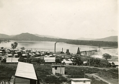 Many small cottages in the front half of the photograph, with a dam in the background, showing scaffolding for construction on the dam wall. Also showing the far bank of the dam with hills in the background. 