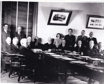 Black and white image of 15 men and 1 women, seated and standing around meeting table. Documents and writing tools on table.