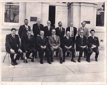 A group of thirteen men in suits out the front of the entrance of a building. Seven men are seated on chairs and the others stand behind.