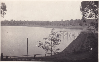A black and white picture of a large lake with a small wooden jetty. There is a road going around the lake. On the other side of the lake there is a line of trees.