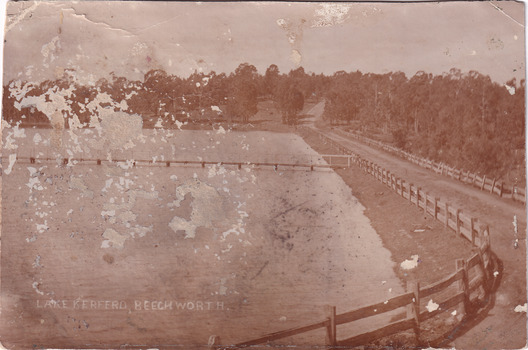 Black and white image of a lake with a wooden jetty. There is a road alongside the lake. This road disappears into a forest of trees on the other side of the lake. 