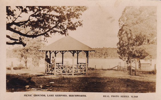 Photograph of a wooden pavilion in front of a large lake. There is a man and women posing against the pavilion. Branches from a large tree can be seen at the top of the image. 