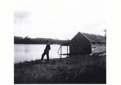 Black and white photograph of a man standing by a large lake. There is a large wooden building behind him.