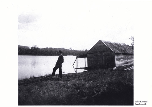 Black and white photograph of a man standing by a large lake. There is a large wooden building behind him.