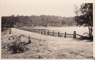 Sepia photograph of the narrow road adjacent to the lake that slightly bends. Trees line the bank in the background and a faint silhouette of the jetty can be seen.