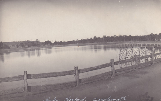 Black and white photograph of still Lake Kerferd, Beechworth. Trees line the bank in the background. Slightly bent horizon with wooden fence.