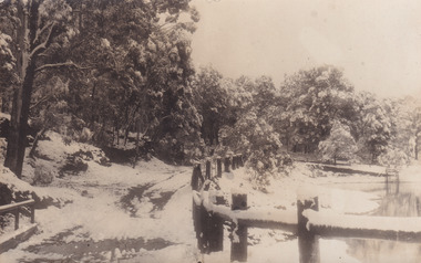 Black and white photograph of a lake and forestry, with snow covering most surfaces of trees, the road and fencing that surrounds the lakes.