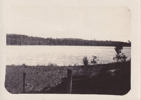 Black and white photograph of a lake, with wire fencing in the fore ground of the photo and this forestry in the far background.  