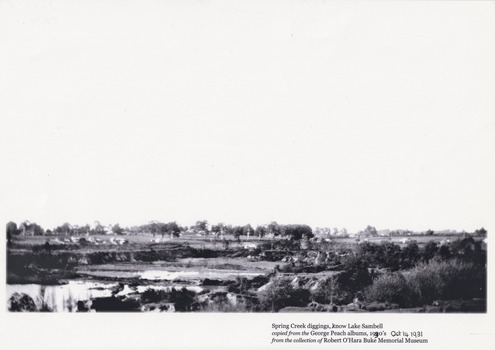 View of Lake Sambell while it was under construction, there is shrubbery in the foreground, and in the back there are rows of trees.