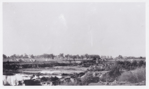 View of Lake Sambell while it was under construction, there is shrubbery in the foreground, and in the back there are rows of trees.