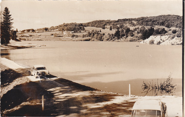 Sepia photograph of Lake Sambell. Trees align on the banks on the background. Two cars seen on the road side.