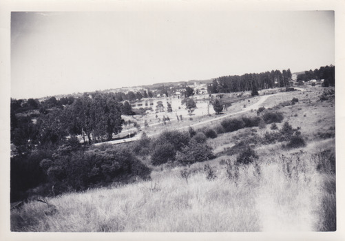 Black and white photograph of a lake and surrounding landscape. Perspective is from a slight incline with shrubs and lightly coloured grass in the foreground, leading into a midground featuring roads, powerlines, with some areas of densely clustered trees. In the centre is a body of water with a divided smaller section at the front. In the background are houses, other buildings, dense trees, and hills. 
