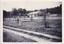 Black and white photograph of camping grounds and building. Dirt road in foreground crosses length of photograph. Grassy campgrounds featuring small trees encased in protective fencing, a barrel drum with the letter ‘B’, and caravan sites with power outlets. Another dirt road runs along a single-story gable roofed white building. Building has four small windows on the length side and two large windows on the width side. Text on the banner hanging on the building is partially visible with the letters ‘RAVAN PARK’. There is a free-standing message board outside the front of the building and a car to the rear right.  Dense trees along background of photograph.