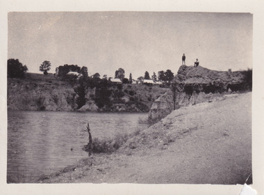 Men standing and sitting on outcrop of rocks at Lake Sambell, with houses in the background. 