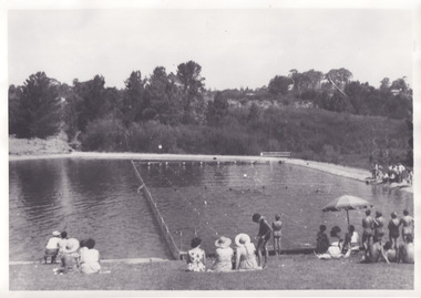 Black and white photograph of people sitting on the grass by a lake watching a swimming carnival.