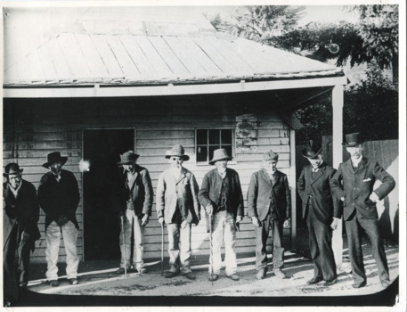 Taken c1900, six elderly Chinese men and two local Beechworth men stand in a row outside a  weatherboard miners hut. The Chinese men wear trousers and jackets with hats or caps, three lean on walking sticks. The two men on the right wear black suits and top hats. The man second from the right is believed to be a Chinese Presbyterian minister.