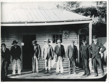 Taken c1900, six elderly Chinese men and two local Beechworth men stand in a row outside a  weatherboard miners hut. The Chinese men wear trousers and jackets with hats or caps, three lean on walking sticks. The two men on the right wear black suits and top hats. The man second from the right is believed to be a Chinese Presbyterian minister.