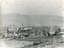 Landscape photograph of a settlement with wooden shack style houses in the foreground, surrounded by worked farm plots. There are industrial style buildings in the background 