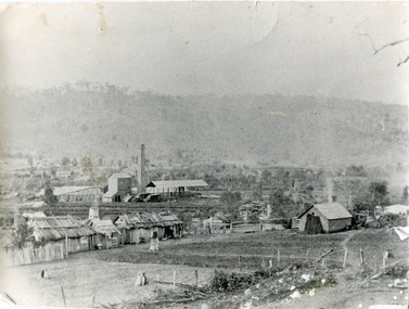 Landscape photograph of a settlement with wooden shack style houses in the foreground, surrounded by worked farm plots. There are industrial style buildings in the background 