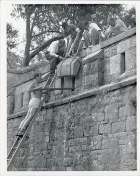 View of outside wall of Powder Magazine Beechworth with a group of workmen at the top and single man poised on a ladder leaning against the wall.