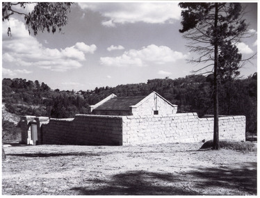 Black and white photo of stone building on a grassy hill with a tree next to it.